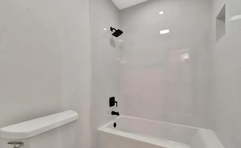 Tub and shower combination with matte black fixtures, recessed shelf, and light-toned tile surround
