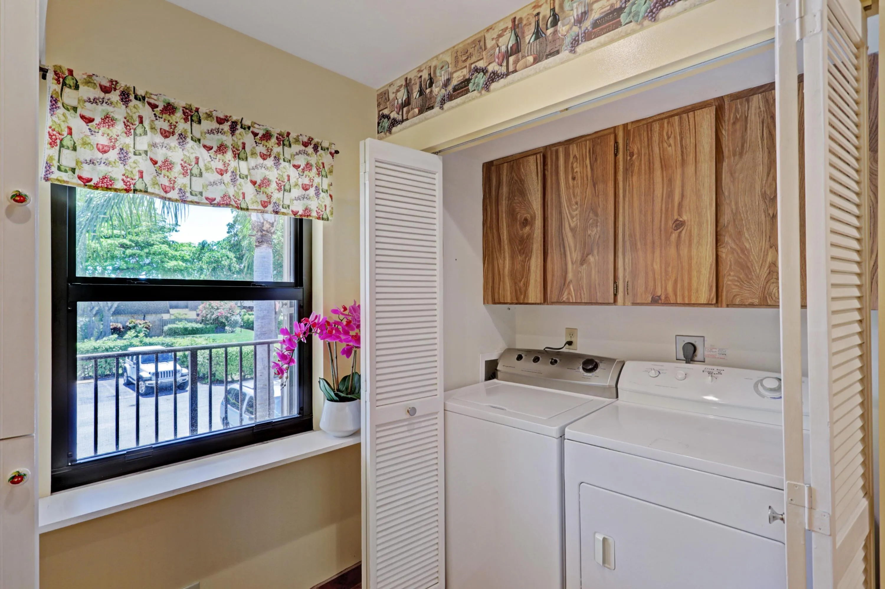 Utility closet featuring wood-finish cabinetry, louvered bi-fold doors, and a window with exterior landscaping visible