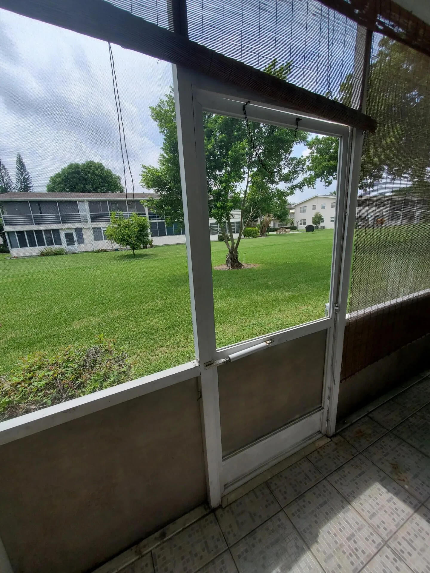 Screened enclosure with tiled flooring and a screen door, overlooking a manicured lawn and mature trees