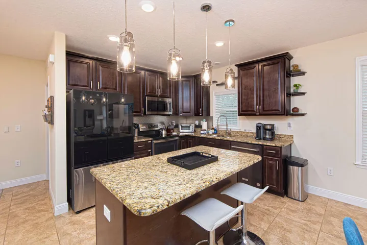 Open-concept kitchen featuring an island with stone-finish countertop, dark wood-finish cabinetry, stainless steel appliances, and pendant lighting