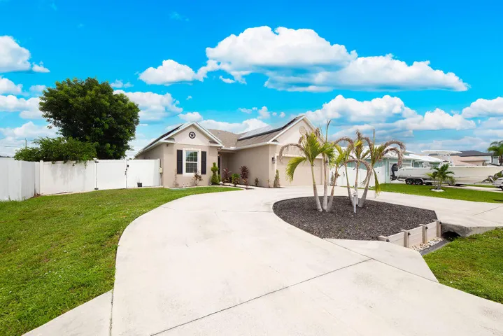Exterior featuring a curved concrete driveway, a central landscape bed with palm trees, a white picket fence, and a pitched roofline
