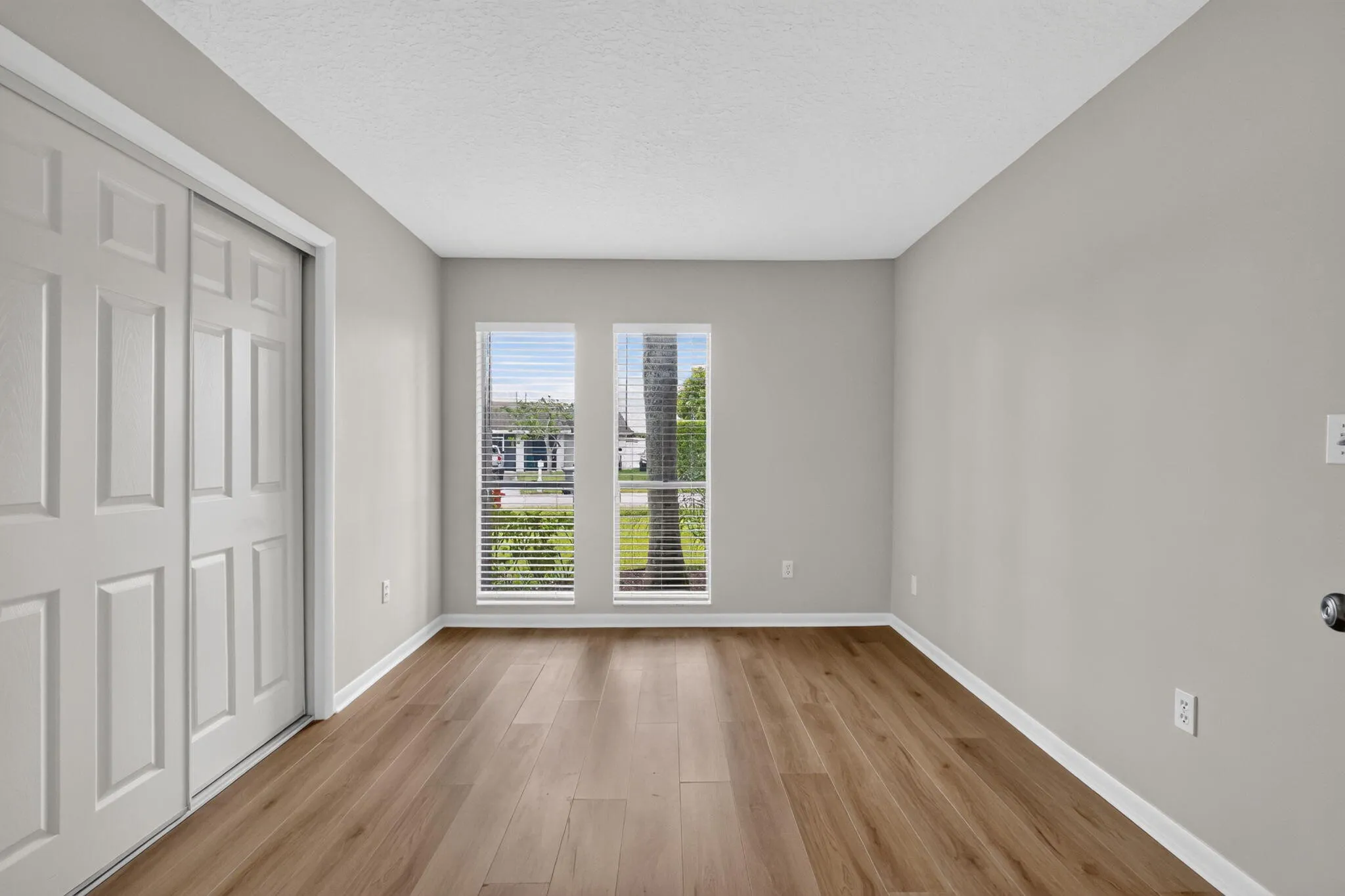 Wood-finish flooring, light grey wall paint, two windows with horizontal blinds, and a bi-fold closet door