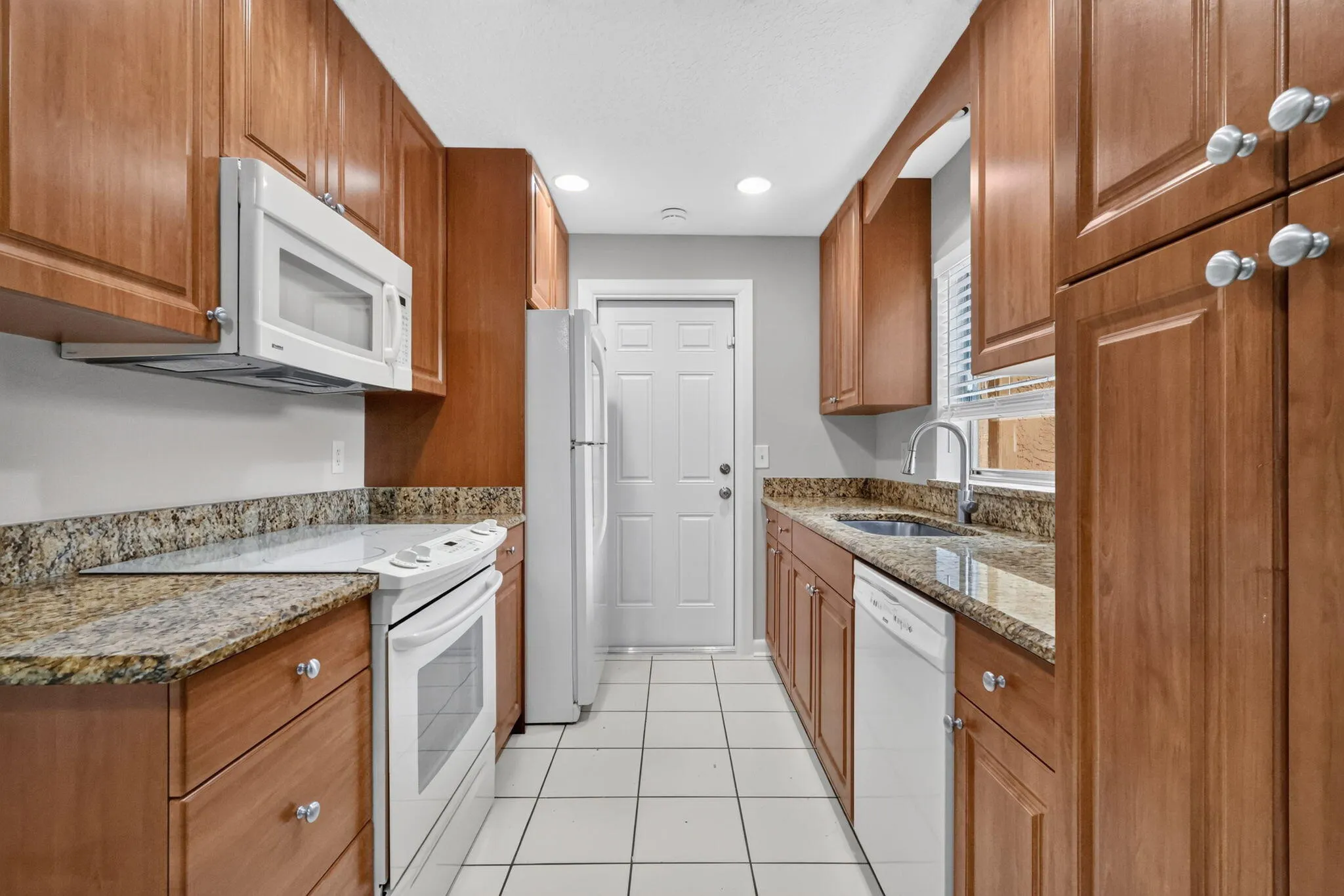 Galley kitchen featuring wood cabinetry, light-toned countertops, white appliances, and white tile flooring