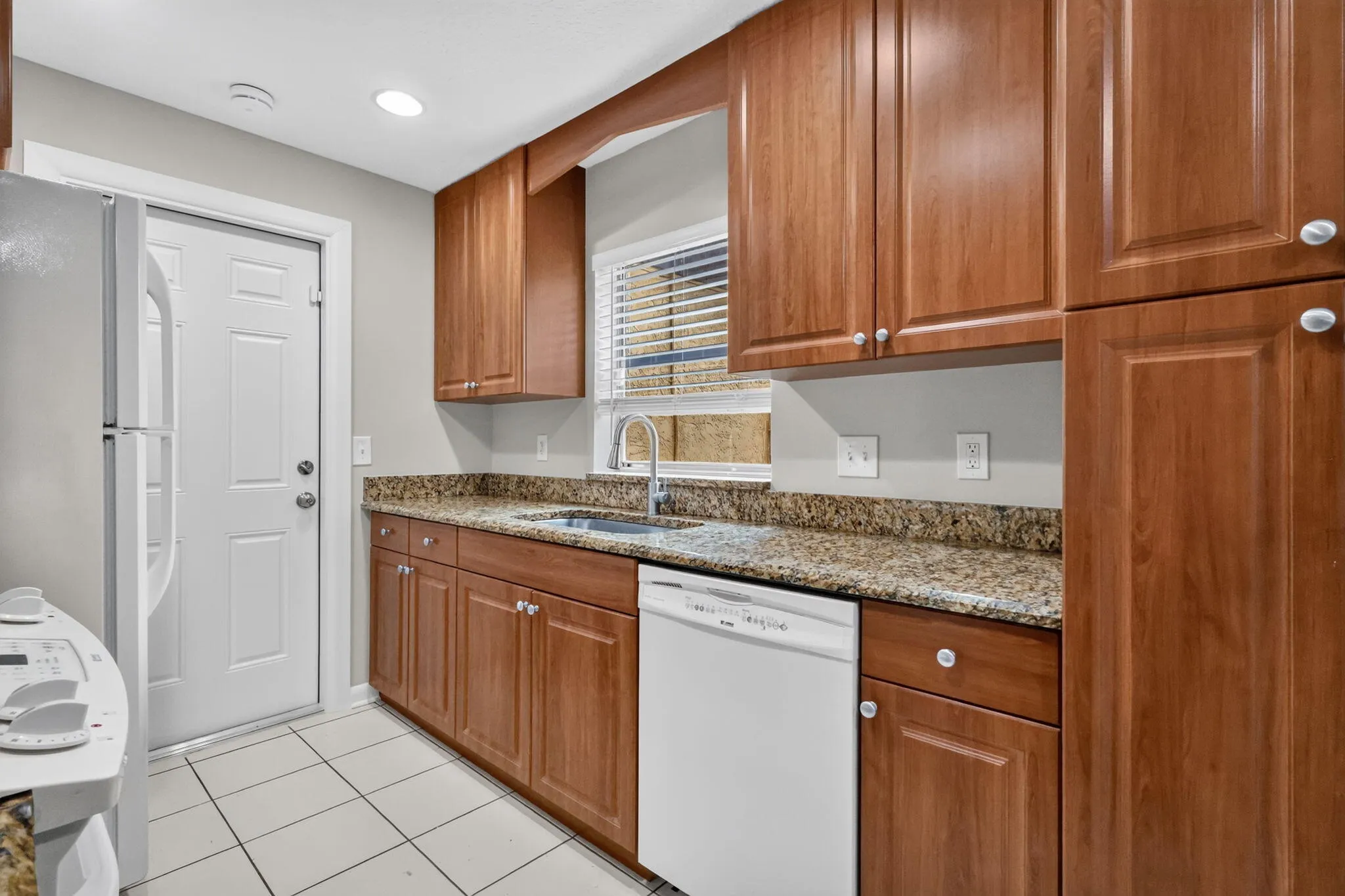 Kitchen featuring wood cabinetry, light-toned countertops, a stainless steel sink with a gooseneck faucet, white appliances, and white tile flooring