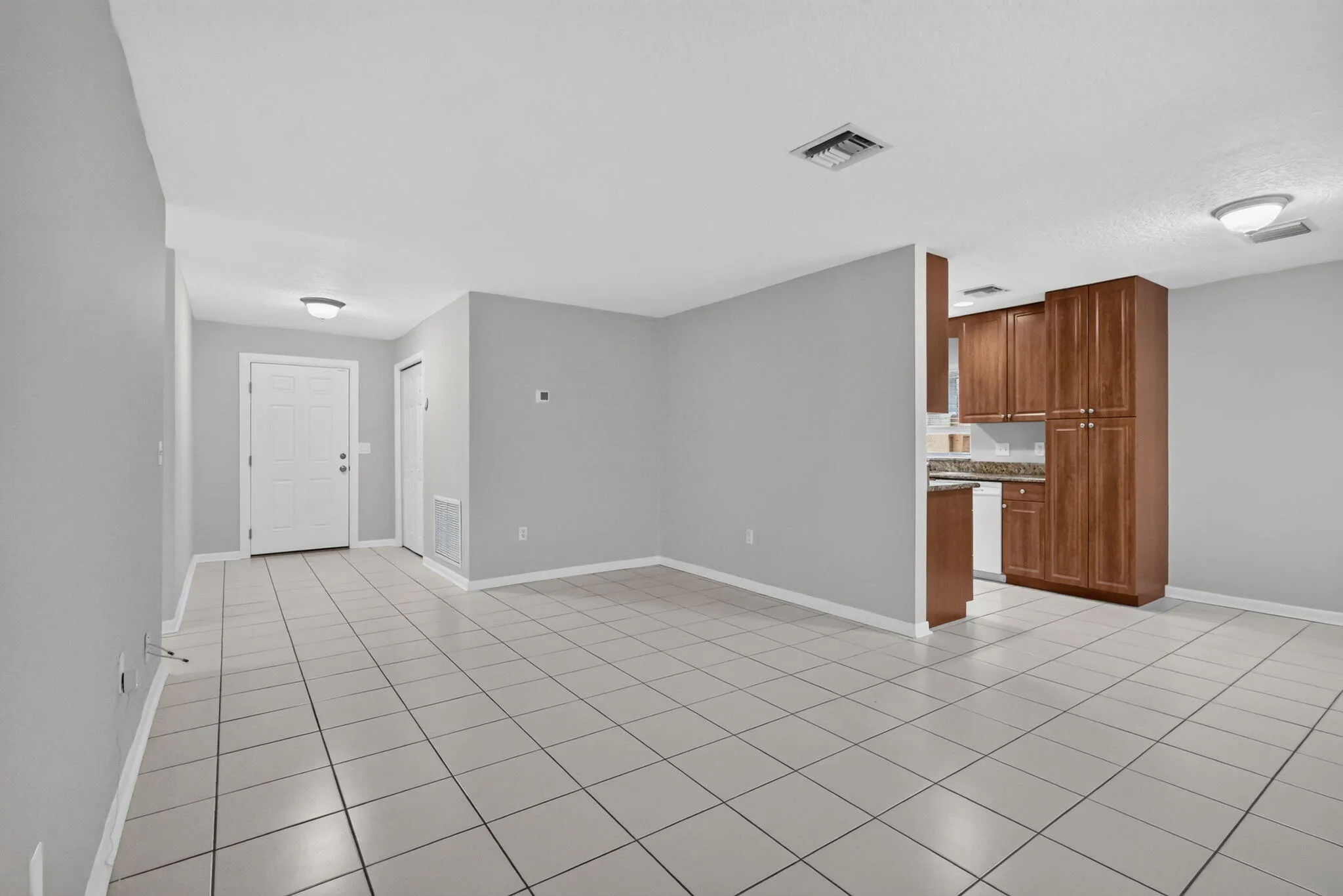 Expansive tiled flooring throughout, complemented by a white paneled entry door and a galley kitchen featuring wood-finish cabinetry and stone countertops
