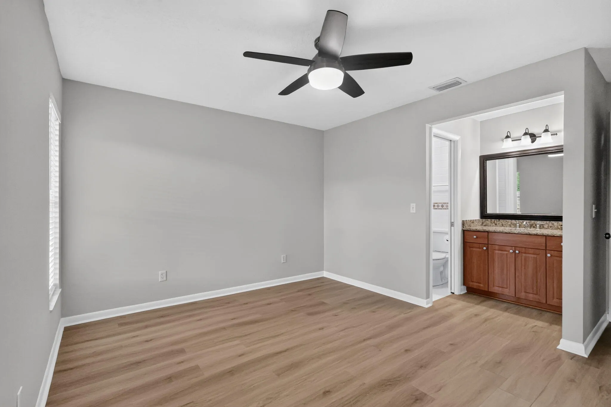 Spacious room featuring wood-finish flooring, a contemporary ceiling fan, and an integrated en-suite bathroom with a wood vanity, granite-style countertop, and mirrored wall cabinet