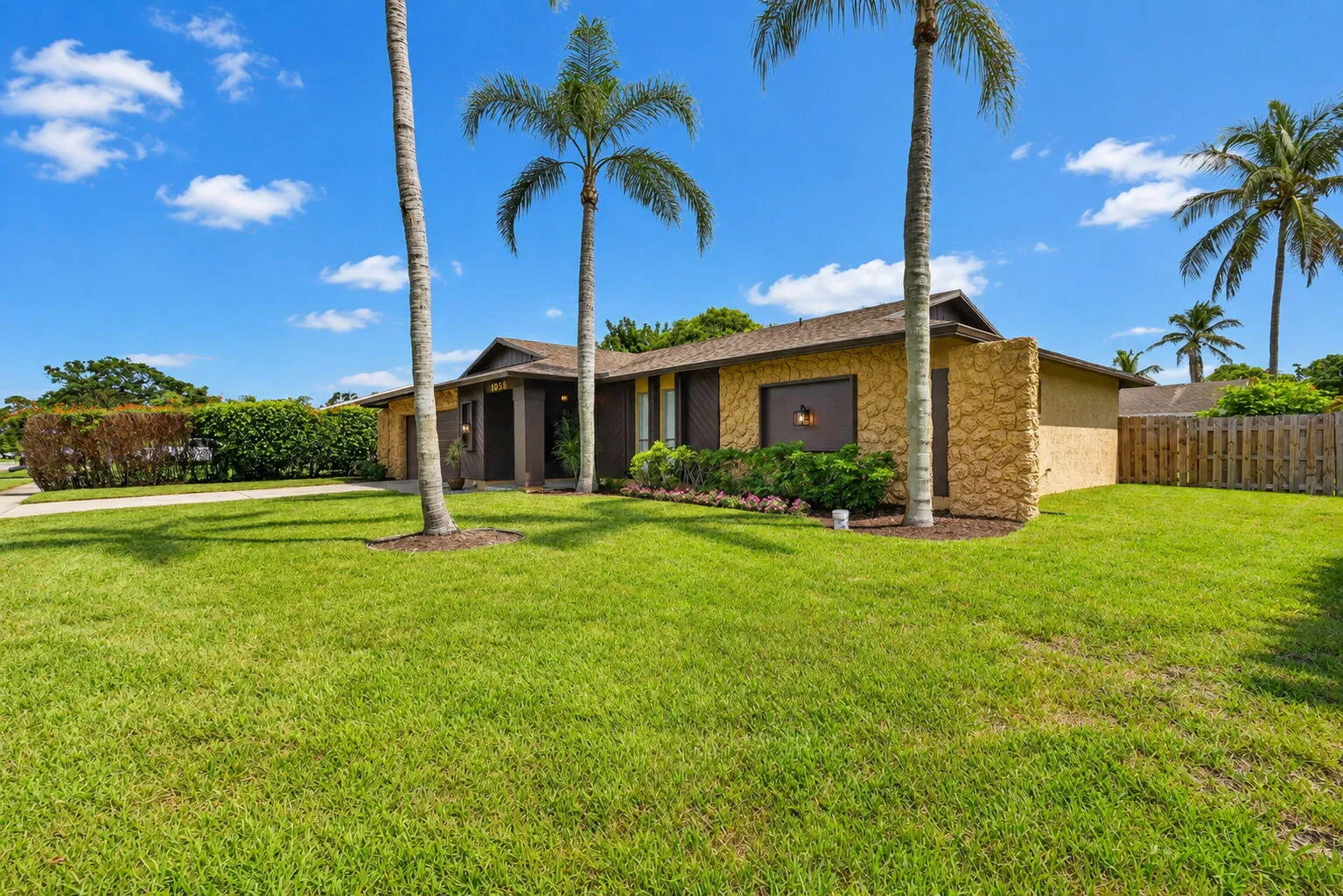 Stone veneer exterior with a wood-shingle roof, featuring established palm trees and a perimeter privacy fence