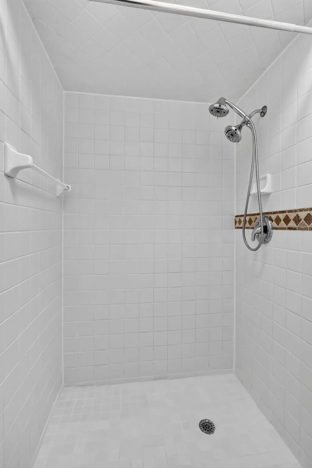 White tiled shower with a decorative mosaic accent band, dual shower heads, and a chrome shower rod