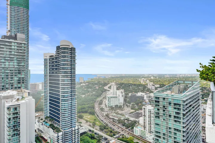 Urban skyline with multiple high-rise buildings, expansive green spaces, a discernible body of water, and a visible transportation corridor