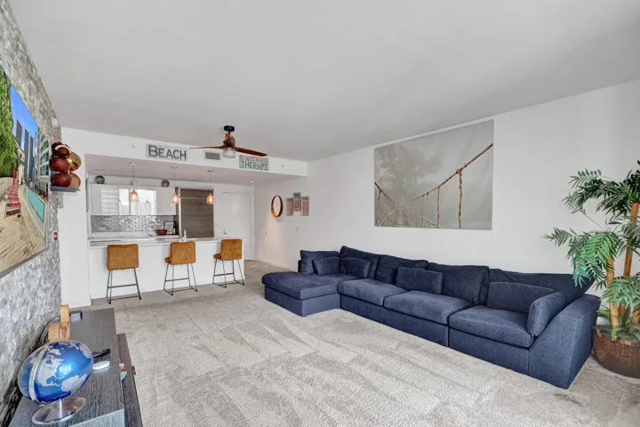 Open-concept living area featuring light-toned carpeting, a kitchen island with pendant lighting, and a ceiling fan with wood-finish blades