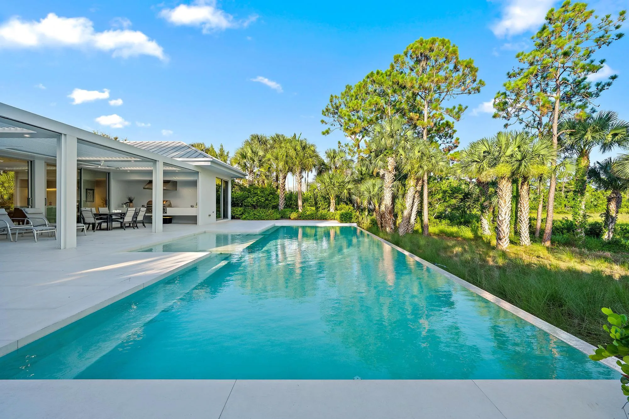 Expansive outdoor area featuring an infinity-edge pool with light-colored coping, a covered patio with a built-in outdoor kitchen, and tropical landscaping