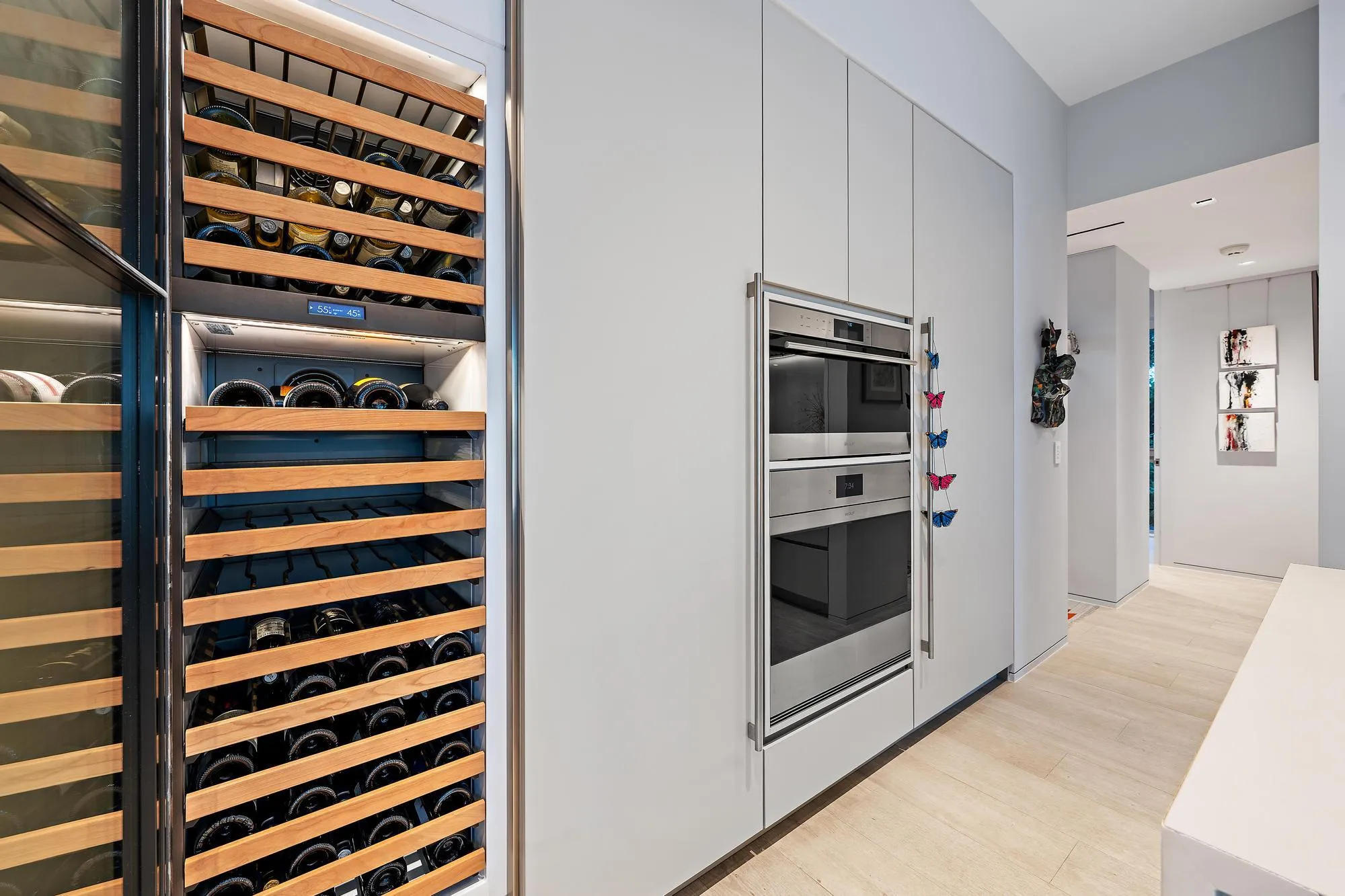 Integrated wine refrigerator with wood racks, built-in double ovens with stainless steel finishes, light wood-finish flooring, and minimalist flat-panel cabinetry