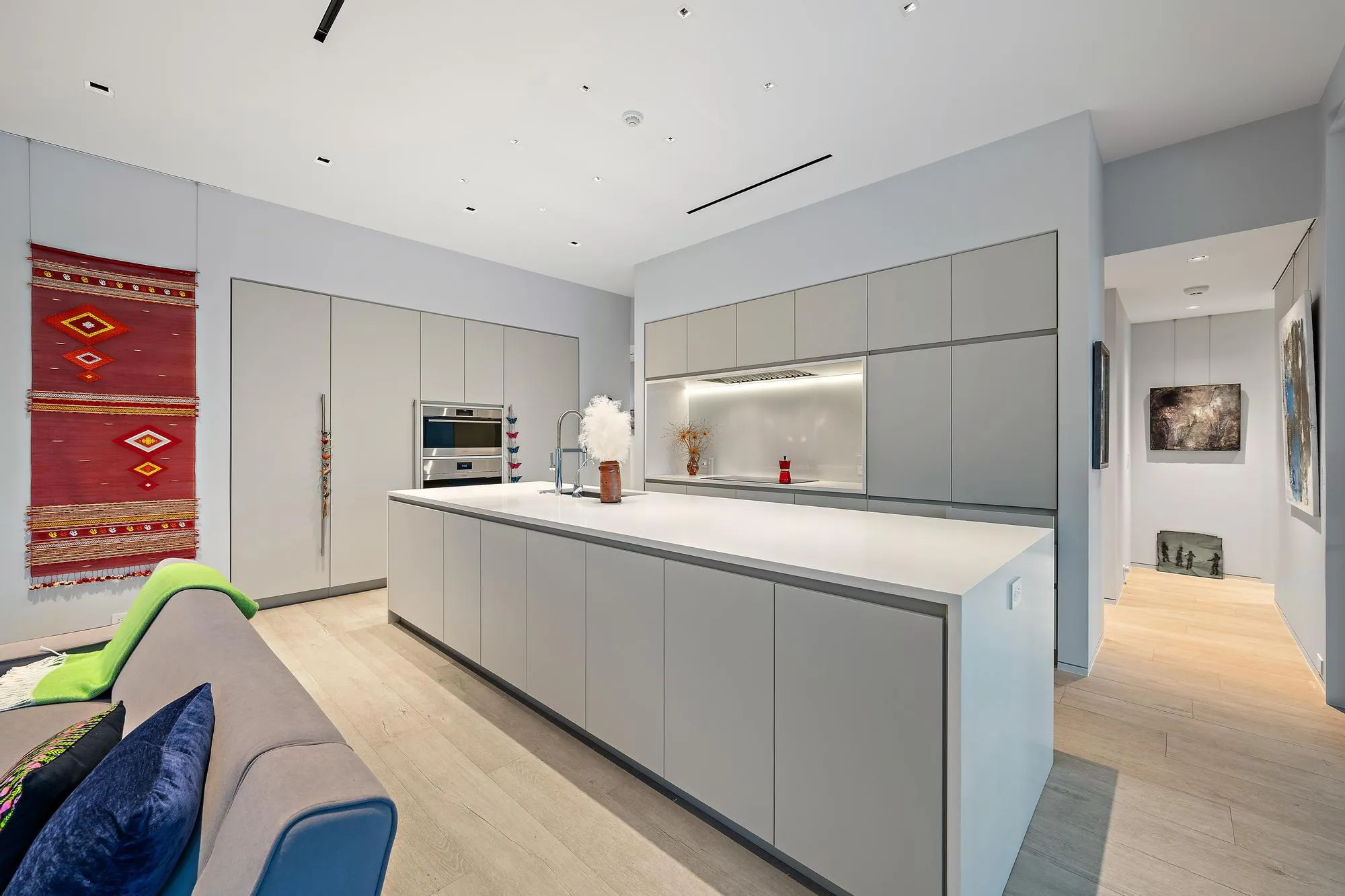 Integrated kitchen featuring a large central island with sink, ample light-grey cabinetry, stainless steel built-in appliances, and light wood-finish flooring