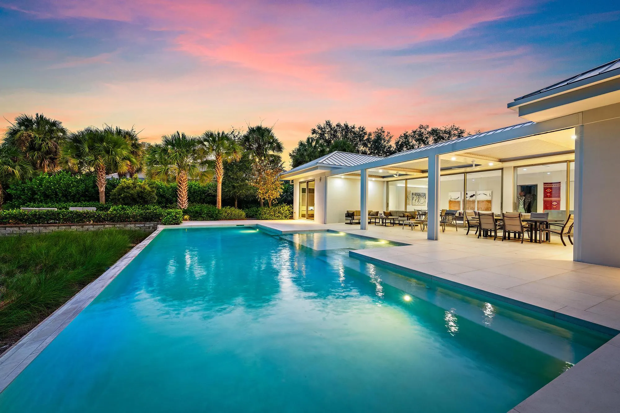 Expansive outdoor living area featuring a rectangular pool with integrated steps, a covered patio, a paver patio, and mature landscaping