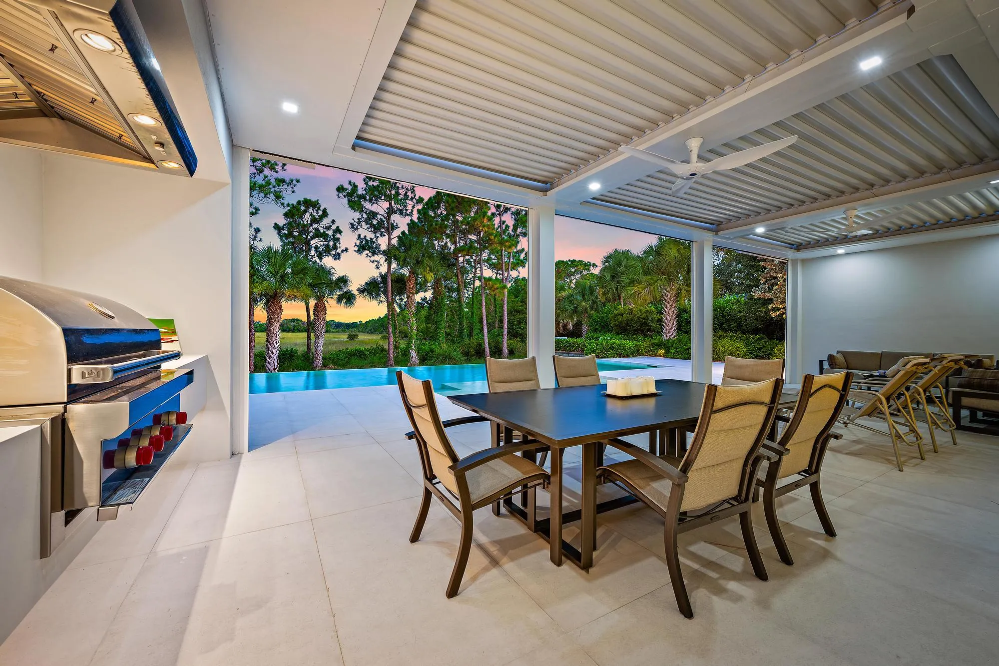 Sophisticated outdoor entertaining space featuring a covered patio with corrugated metal roof and integrated lighting, stone-finish flooring, and a wood-finish dining table with upholstered seating