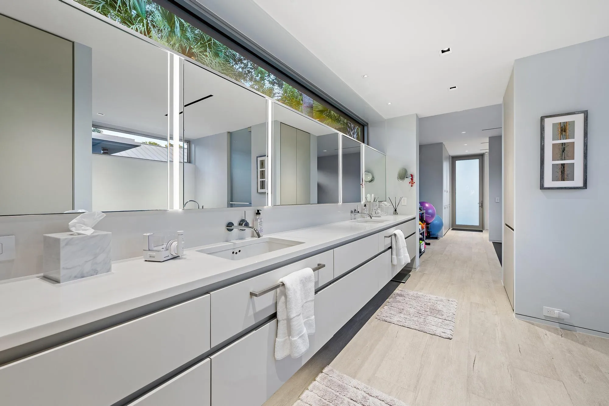Dual vanity with integrated sinks, white countertops, built-in medicine cabinets with integrated lighting, wood-finish flooring, and a large horizontal window