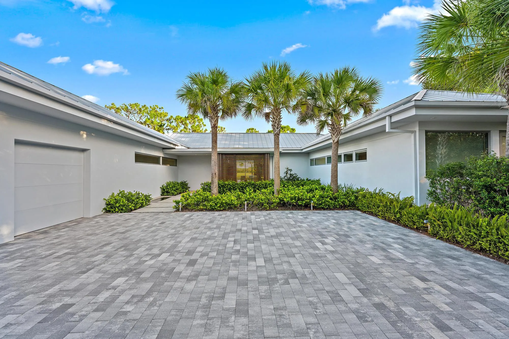 Elegant driveway featuring permeable pavers in a geometric pattern, bordered by lush tropical landscaping with mature palm trees and manicured hedges