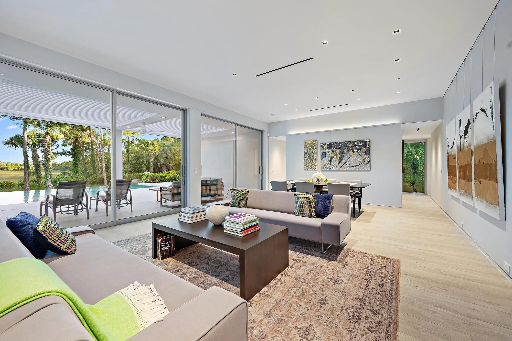 Sophisticated open-concept living space featuring floor-to-ceiling sliding glass doors opening to expansive outdoor grounds, light wood-finish flooring throughout, recessed ceiling lighting, neutral wall finishes, and a dark wood rectangular coffee table anchoring the seating area