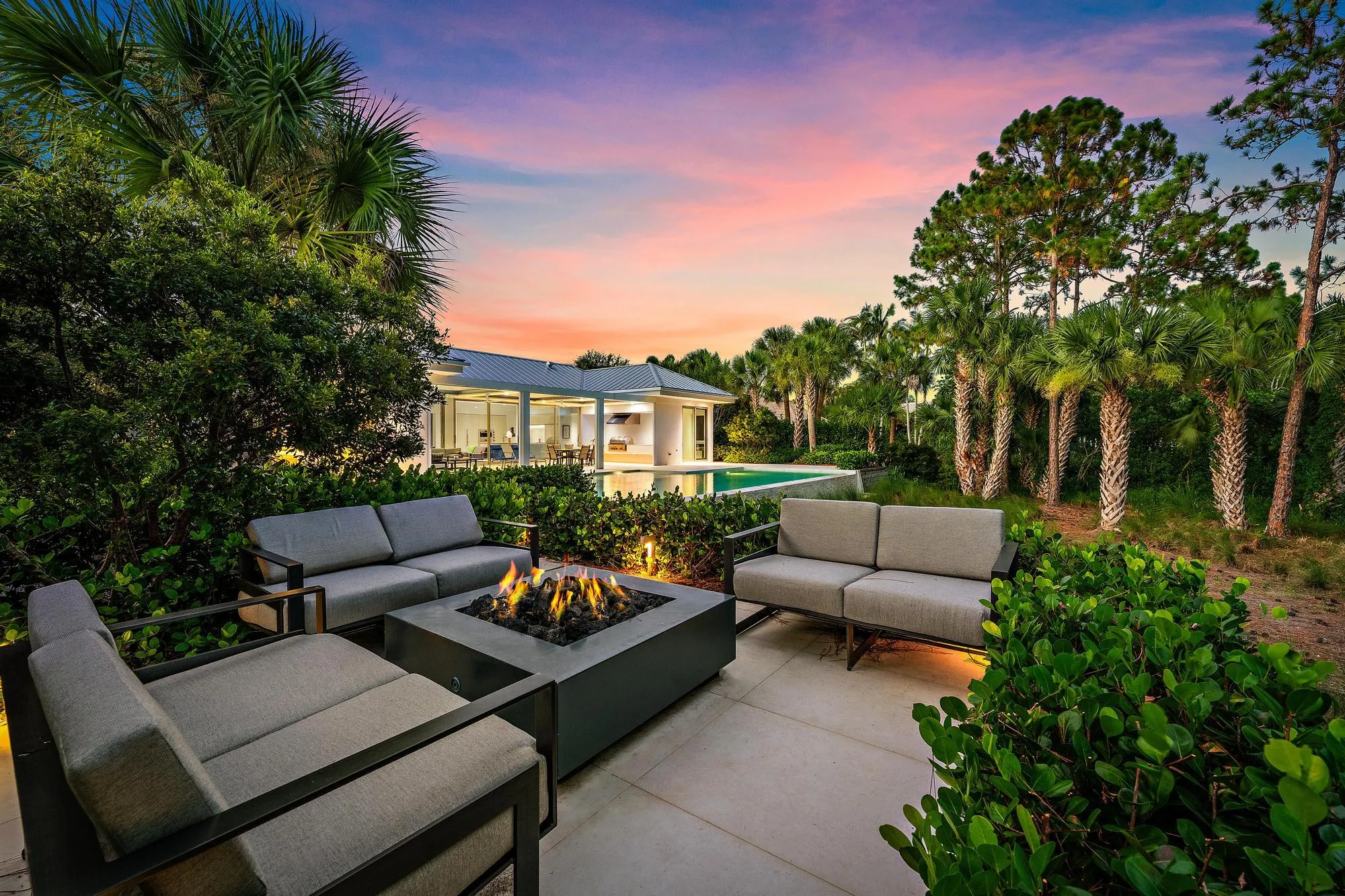 Paver patio with a built-in fire pit, lush landscaping, and mature palm trees