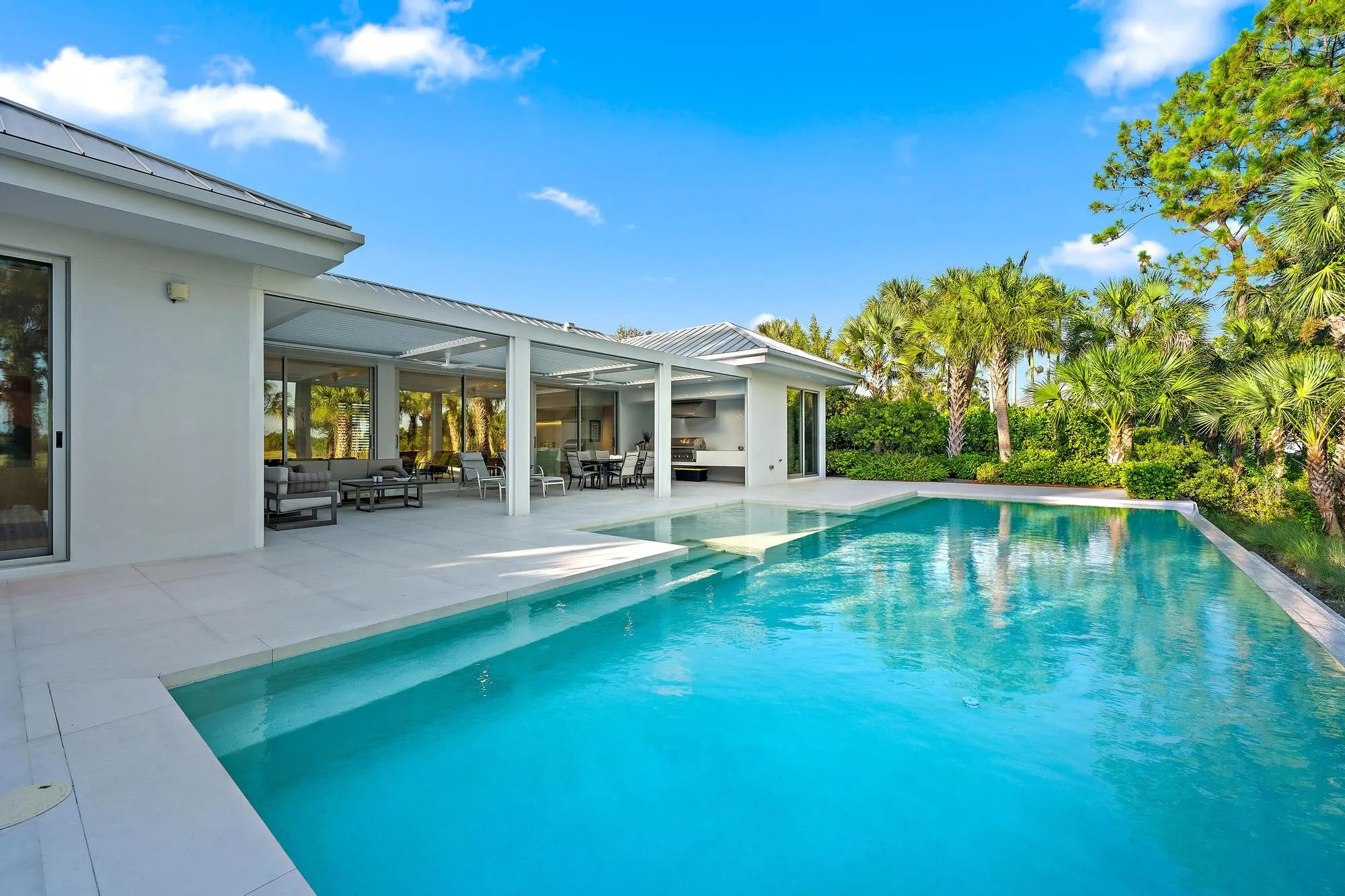 Resort-style saltwater swimming pool with turquoise finish and generous sun deck, anchored by a modern white pergola structure and expansive covered loggia featuring floor-to-ceiling sliding glass doors, mature tropical landscaping with native palms and lush greenery framing the outdoor living space