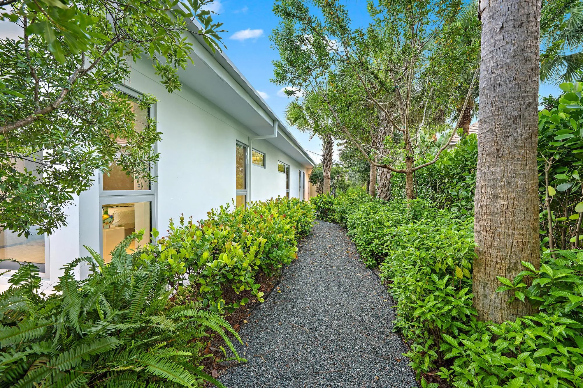 Gravel pathway bordered by abundant green shrubbery, mature trees, and a white exterior wall with multiple windows