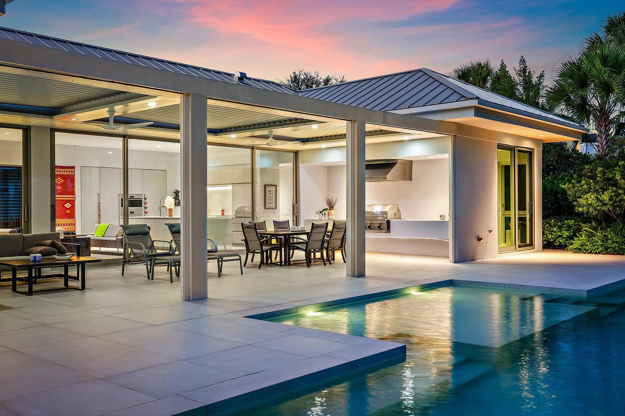 Modern residence featuring a resort-style backyard with an illuminated saltwater pool, expansive travertine-finish patio, and seamless indoor-outdoor living spaces