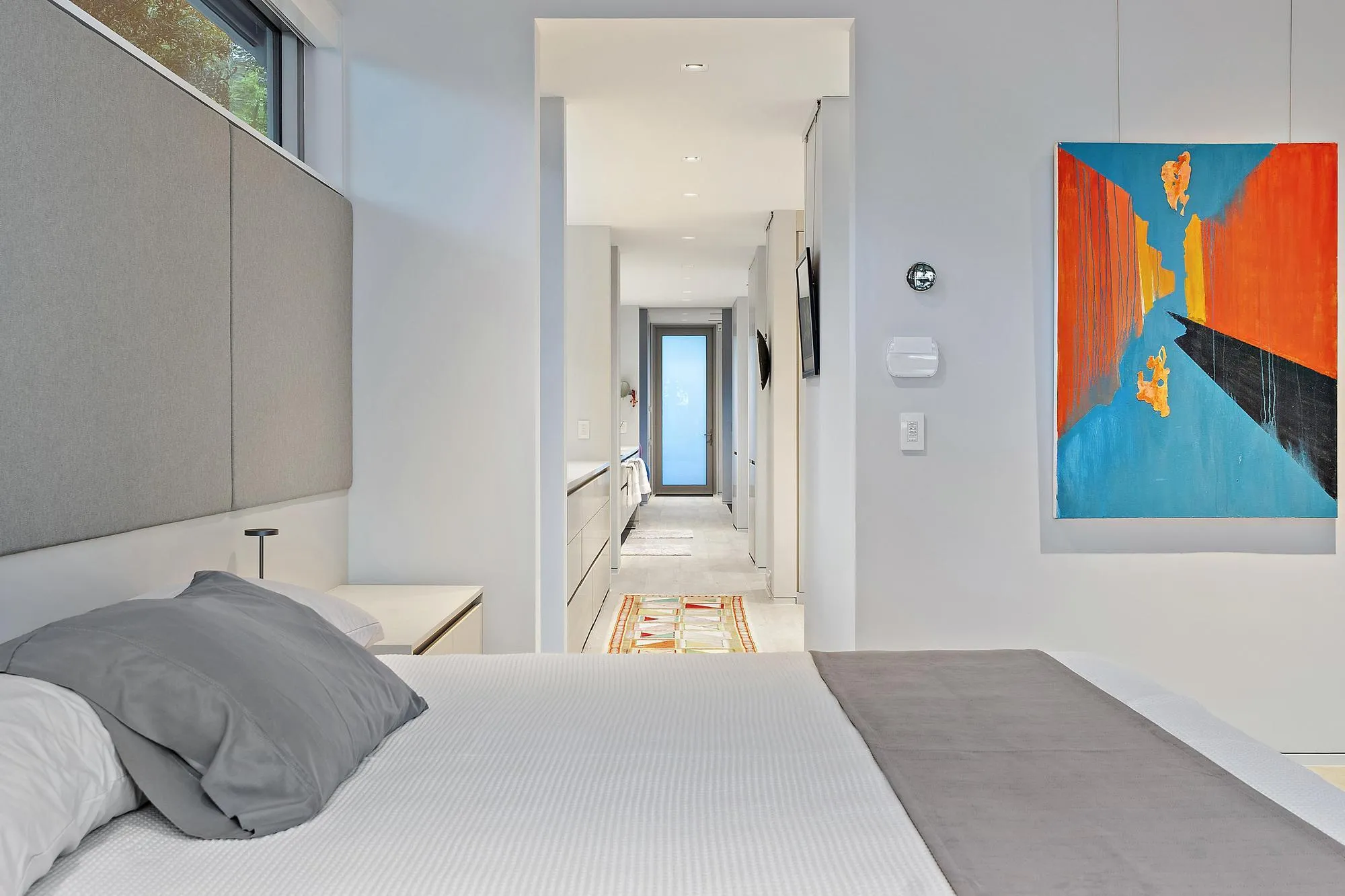 Modern primary bedroom featuring a neutral color palette with light wood-finish flooring, contemporary headboard wall with integrated lighting, and direct hallway access