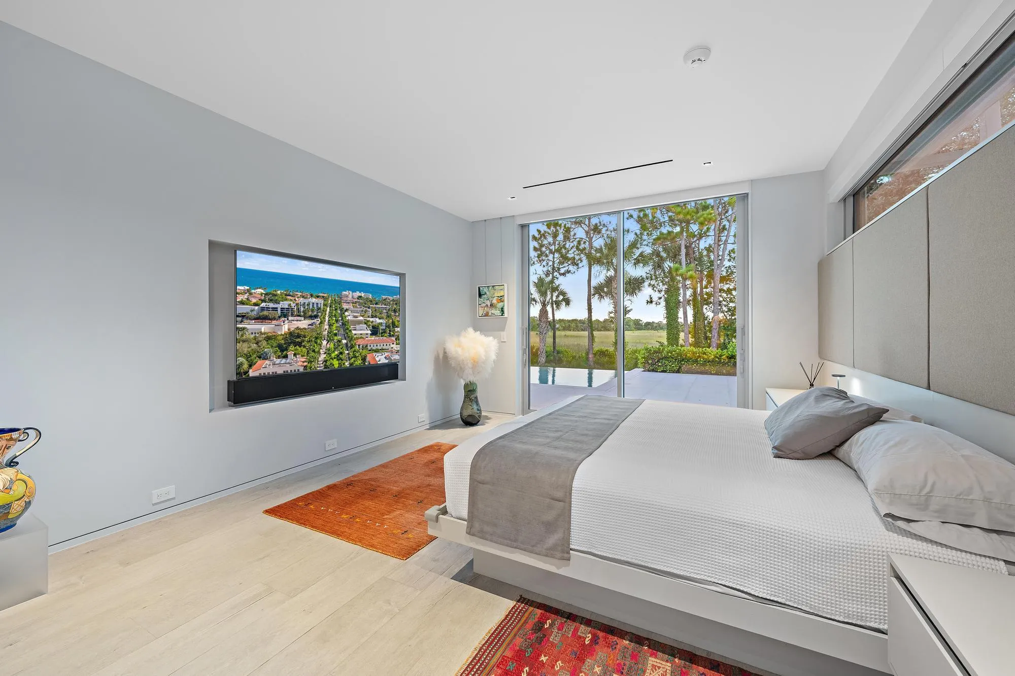 Recessed wall-mounted television, full-height sliding glass doors, built-in headboard, wood-finish flooring, and integrated ceiling lighting