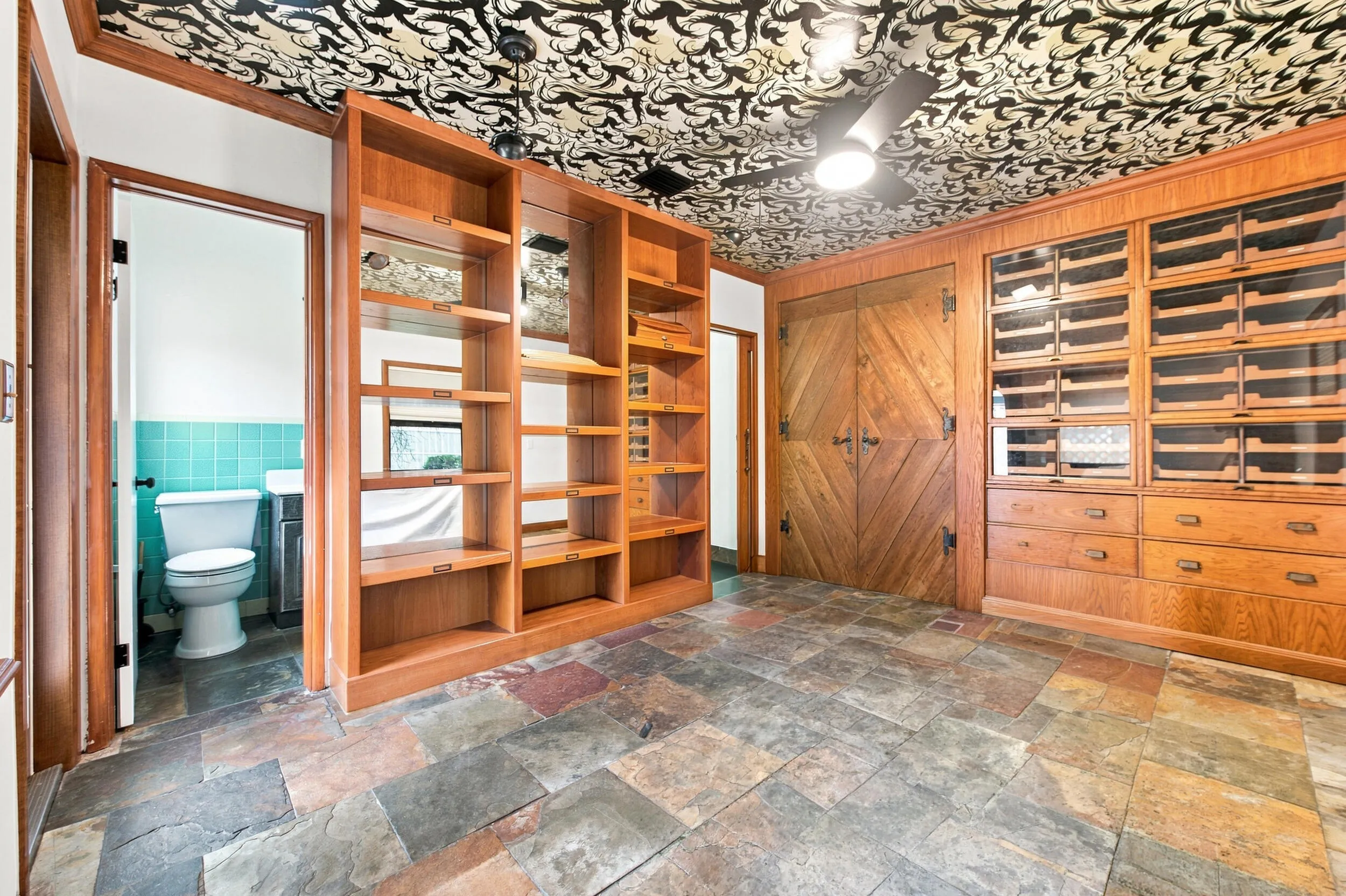 Custom built-in shelving, glass-front cabinetry with pull-out drawers, natural wood double doors, a ceiling fan, and multi-tone natural stone flooring