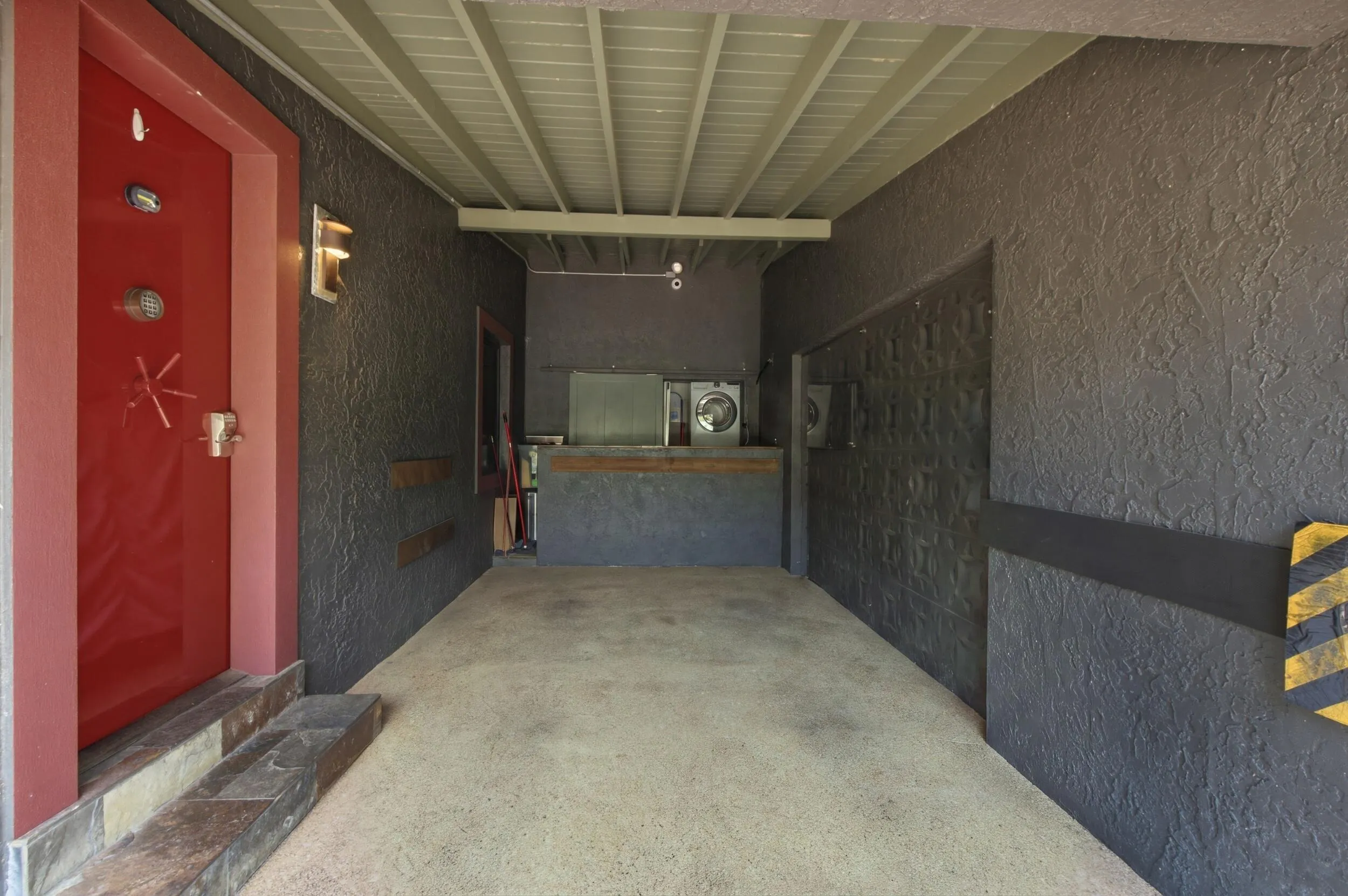 Red entry door with unique hardware, textured exterior wall, built-in laundry appliances, exposed ceiling joists, and concrete flooring