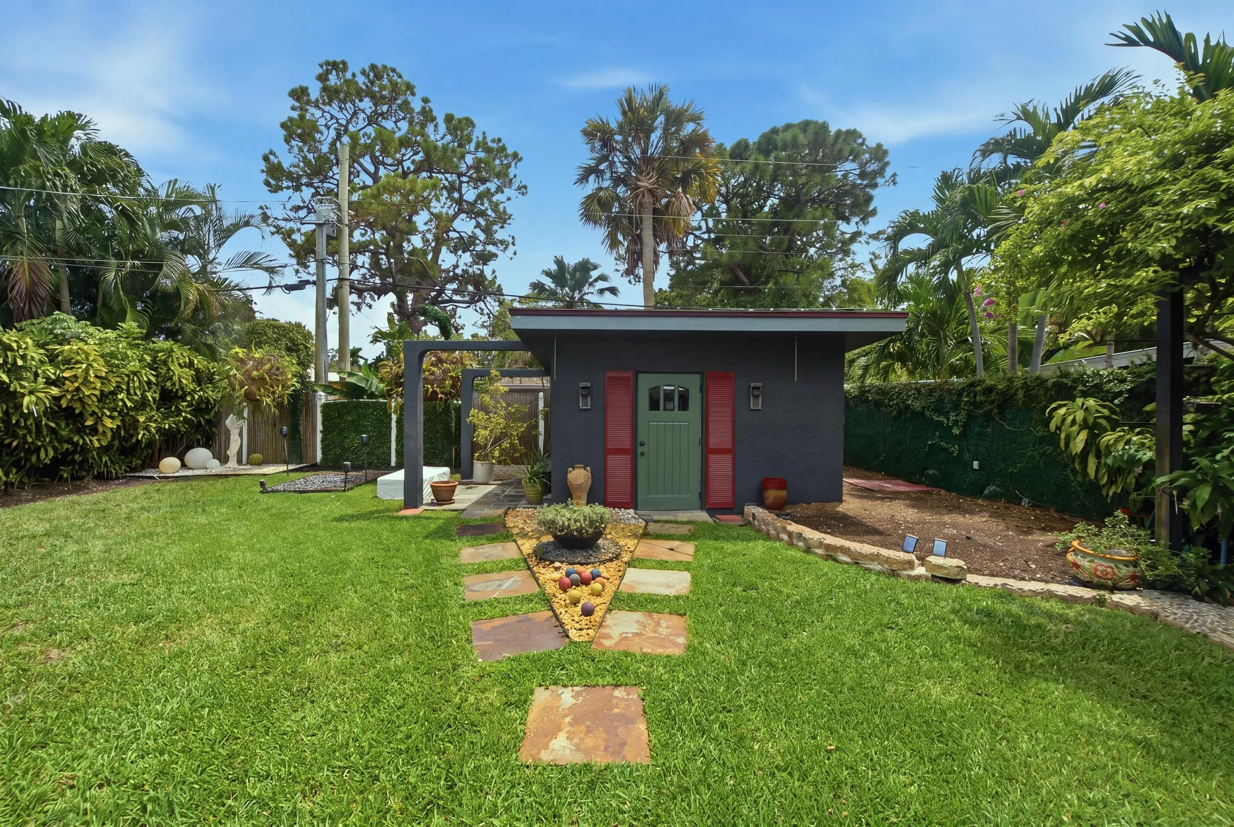 Detached structure with a green door and red shutters, stone-paver pathway, and garden beds