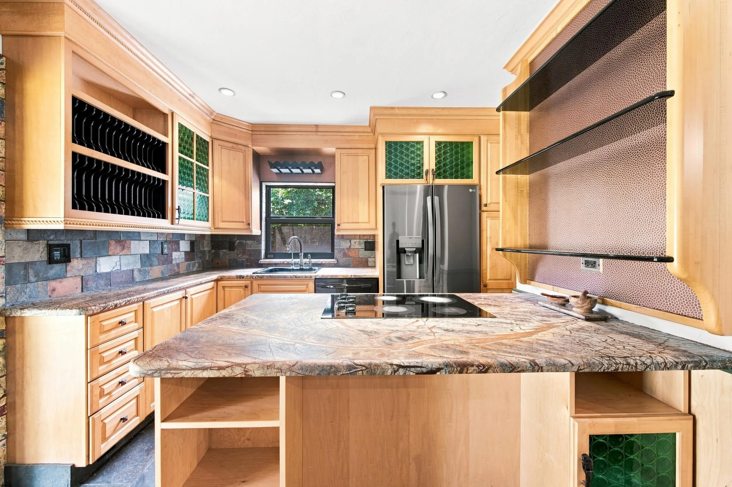 Light wood cabinetry with integrated wine storage, a stone tile backsplash, a kitchen island with a cooktop, built-in shelving, and a stainless steel refrigerator