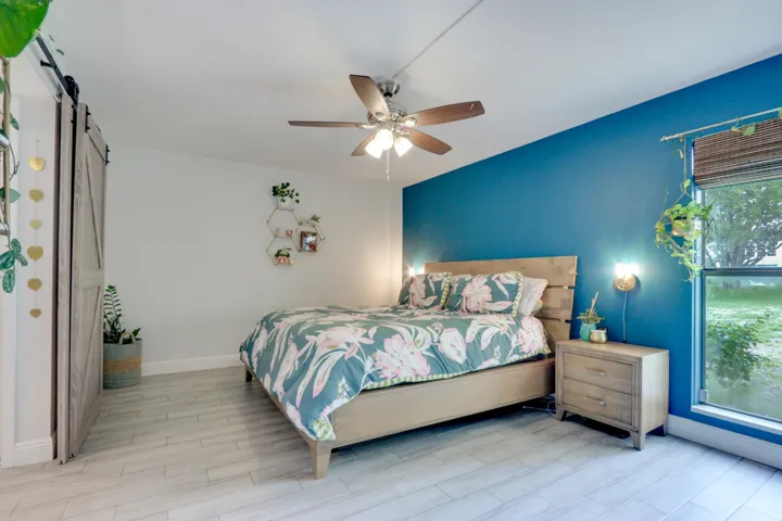 Wood-finish flooring, accent wall, ceiling fan, sliding barn door, and a large window with blinds