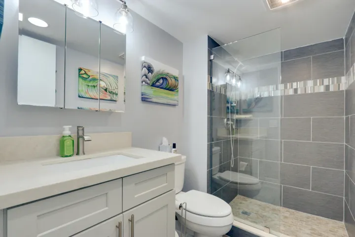 Vanity with a white countertop and undermount sink, mirrored medicine cabinet with integrated lighting, glass-enclosed shower with gray tiled walls, and a mosaic tile accent band