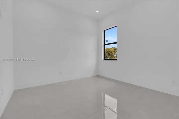 Neutral interior room featuring large-format tile flooring, smooth white walls, recessed ceiling lighting, and a window with a black frame