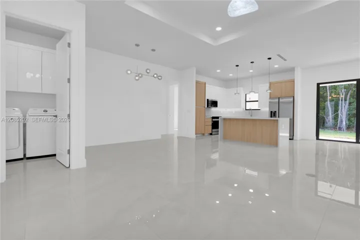 Open-concept interior featuring large-format tile flooring, recessed tray ceiling, kitchen with wood-finish cabinetry, waterfall-edge island, and a dedicated laundry area with built-in storage