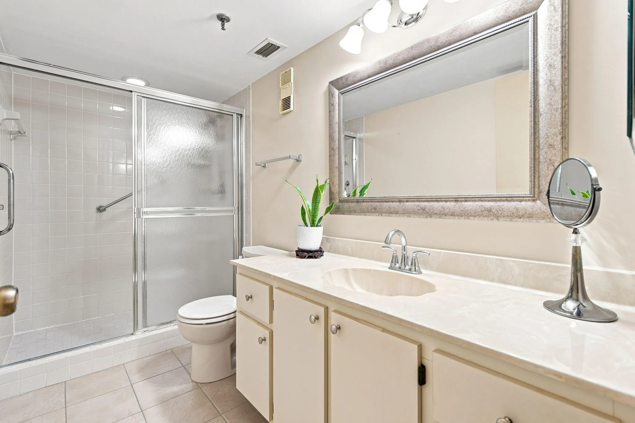 Walk-in shower with clear and frosted glass doors, a vanity with an integrated sink and light-colored countertop, and a large framed mirror with overhead lighting