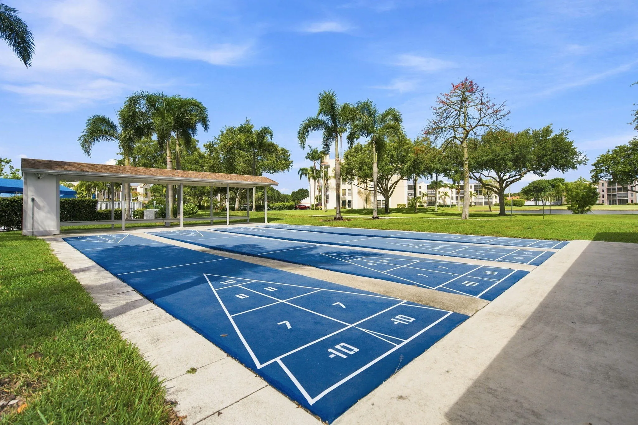 Dual shuffleboard courts with painted markings, covered spectator area, surrounded by green lawns and mature palm trees