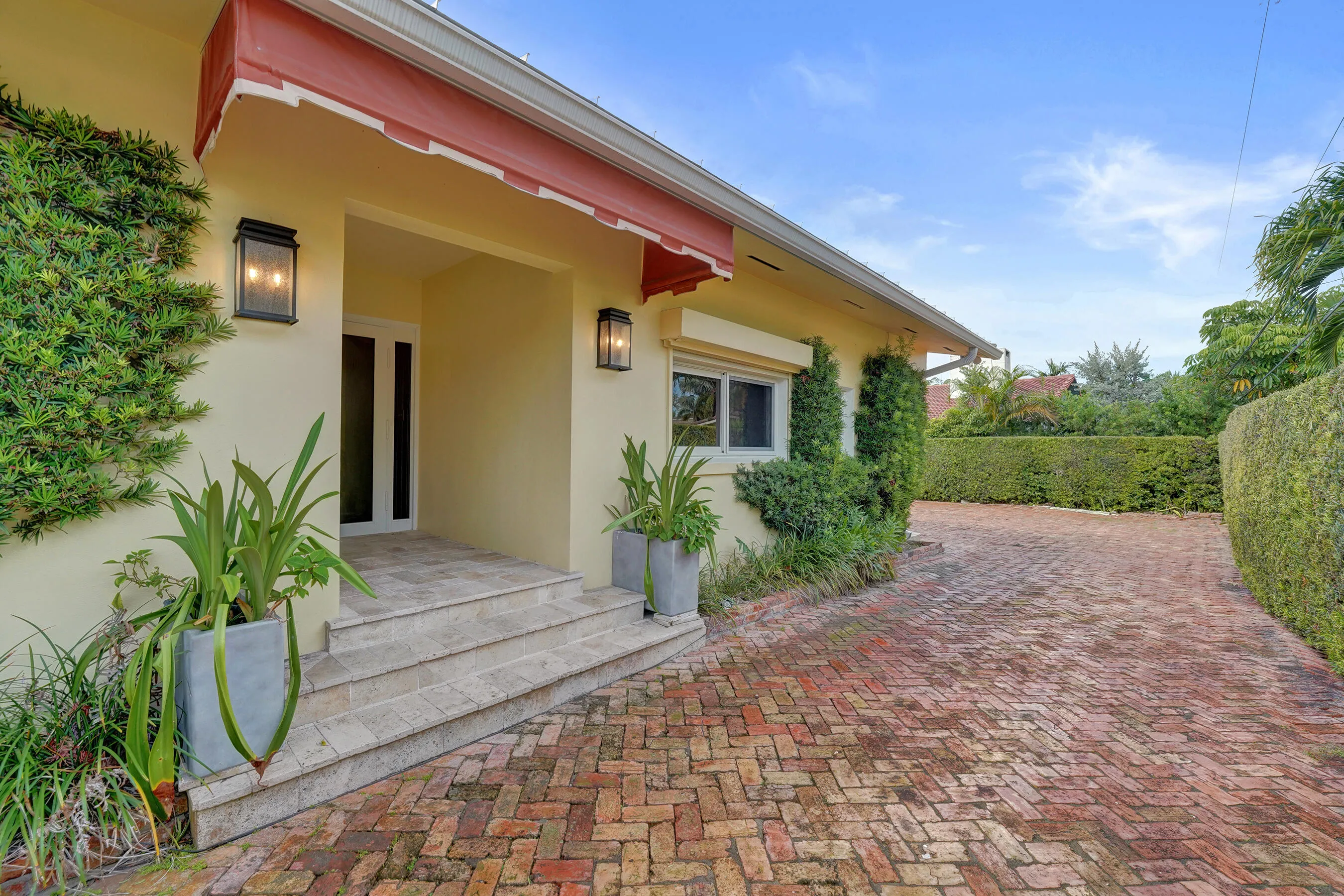 Brick paver driveway, stone entry steps, exterior wall sconces, mature perimeter hedging, and red awning.