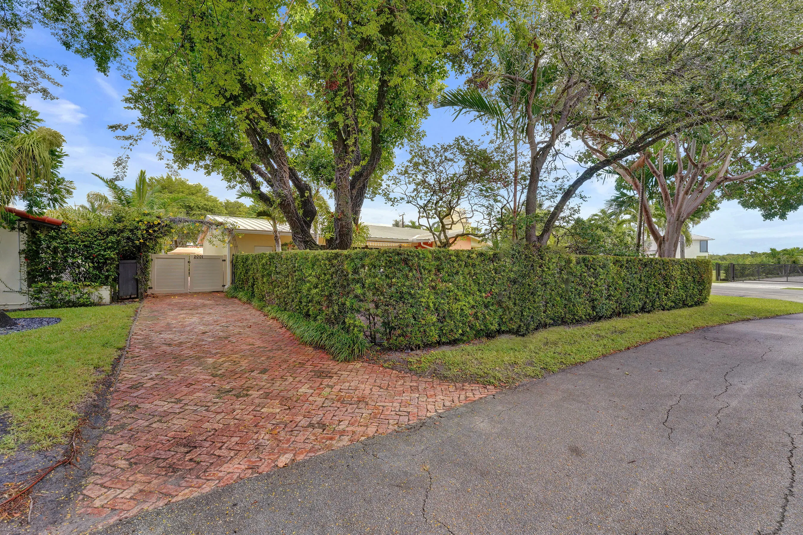Brick paver driveway with a double gate, surrounded by a manicured hedge and mature trees