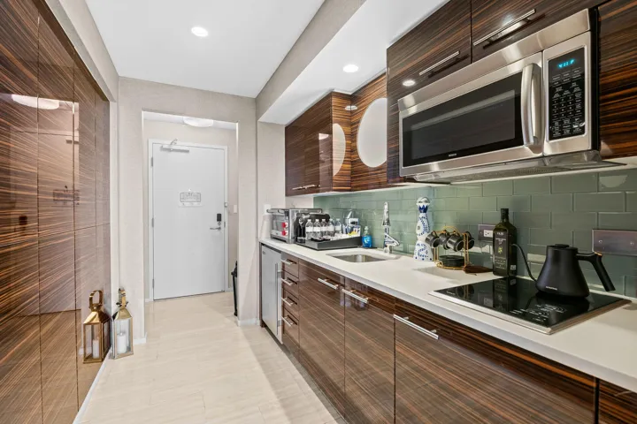 Compact kitchen featuring wood-grain cabinetry, stainless steel microwave, light-colored countertops, tiled backsplash, and an integrated cooktop