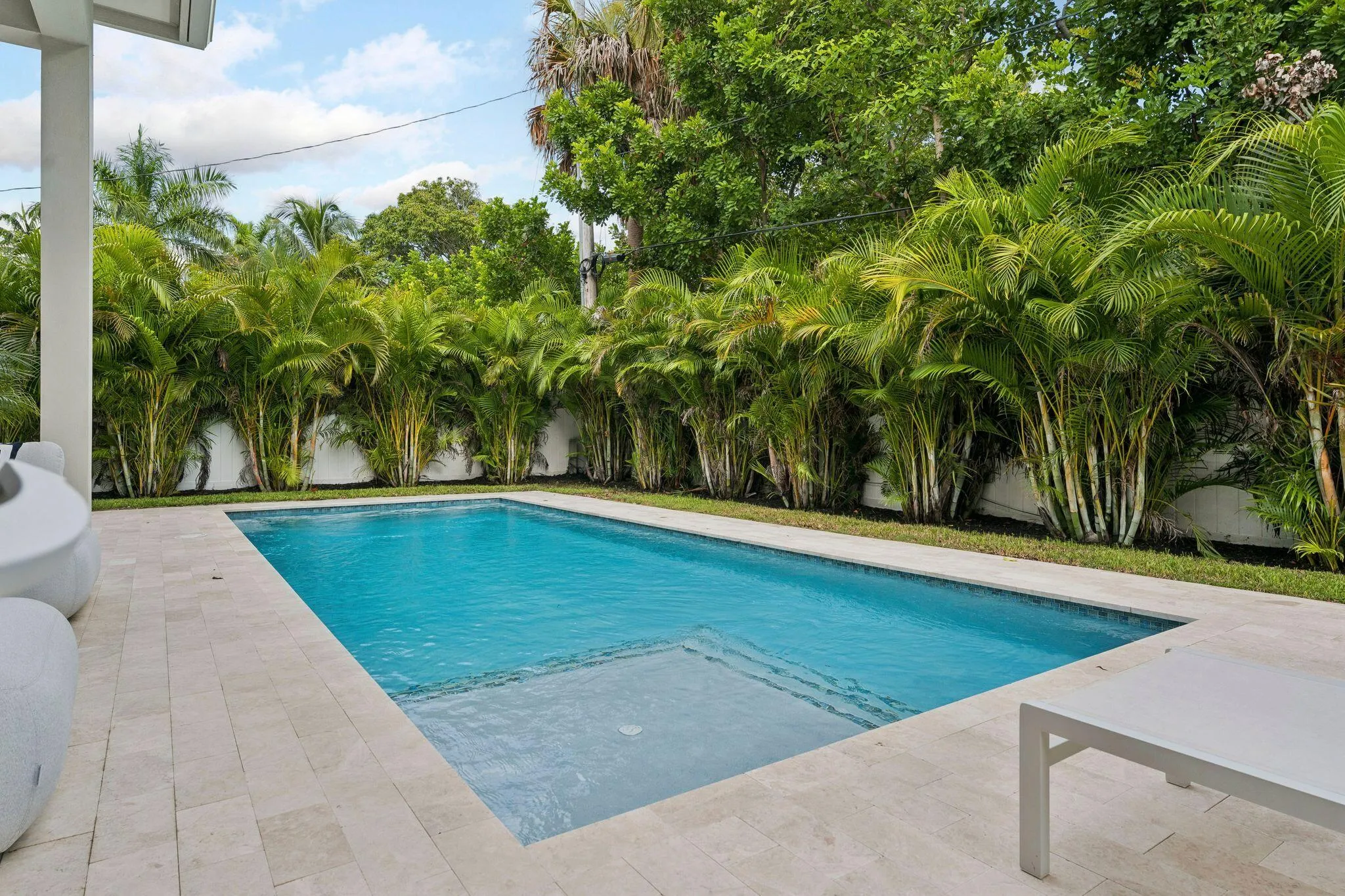 Rectangular swimming pool with integrated steps, surrounded by light-toned paver decking, and lush green landscaping