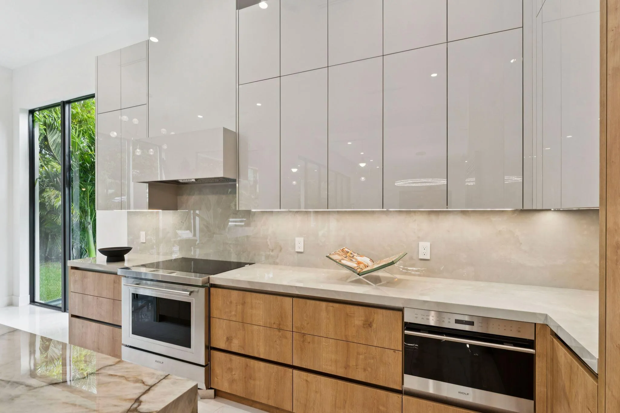 Sleek kitchen featuring light-toned upper cabinetry, wood-finish lower cabinetry, stone countertops, and integrated stainless steel appliances