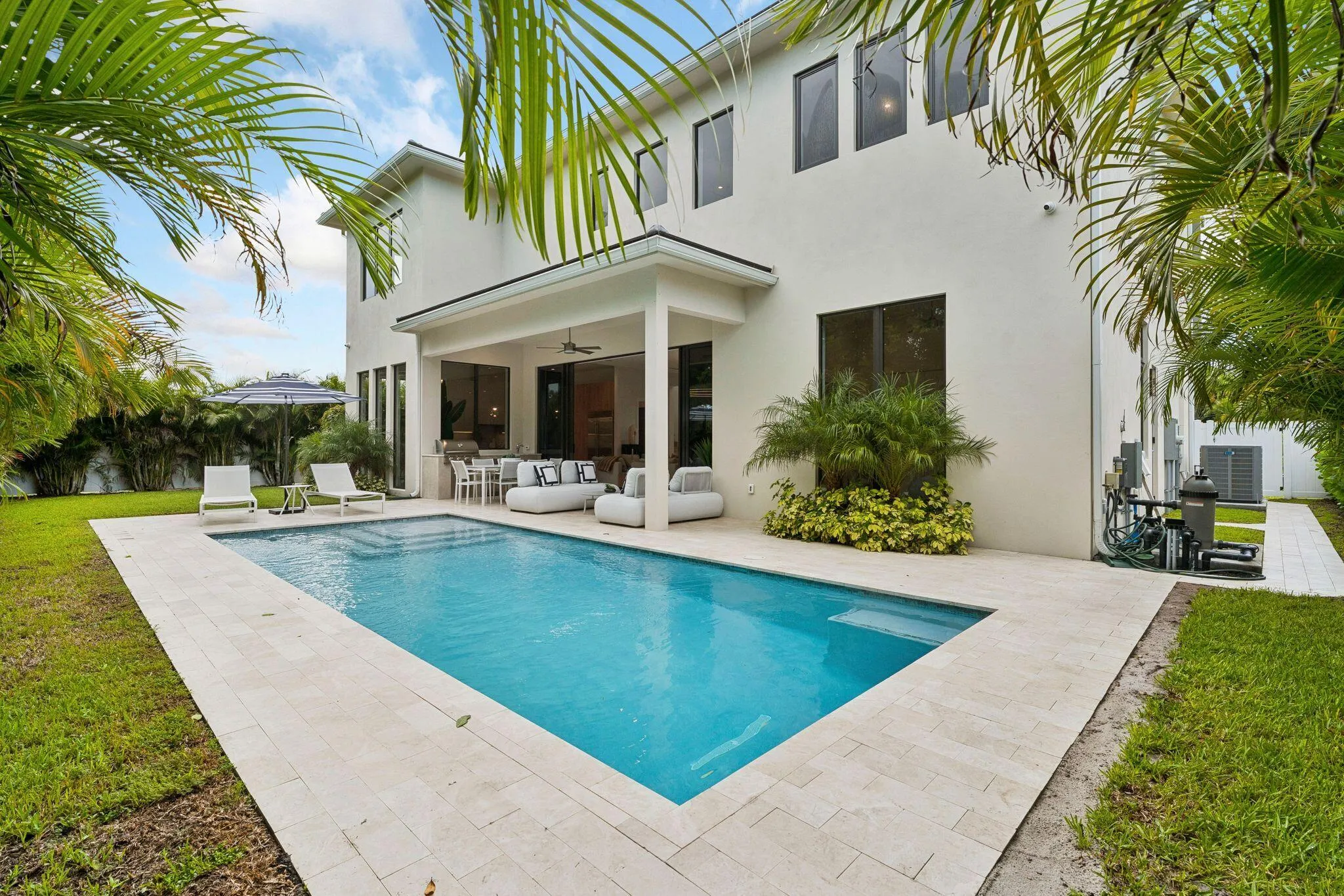 Spacious exterior featuring a rectangular pool, light-toned paver surround, covered patio with recessed lighting, and lush landscaping