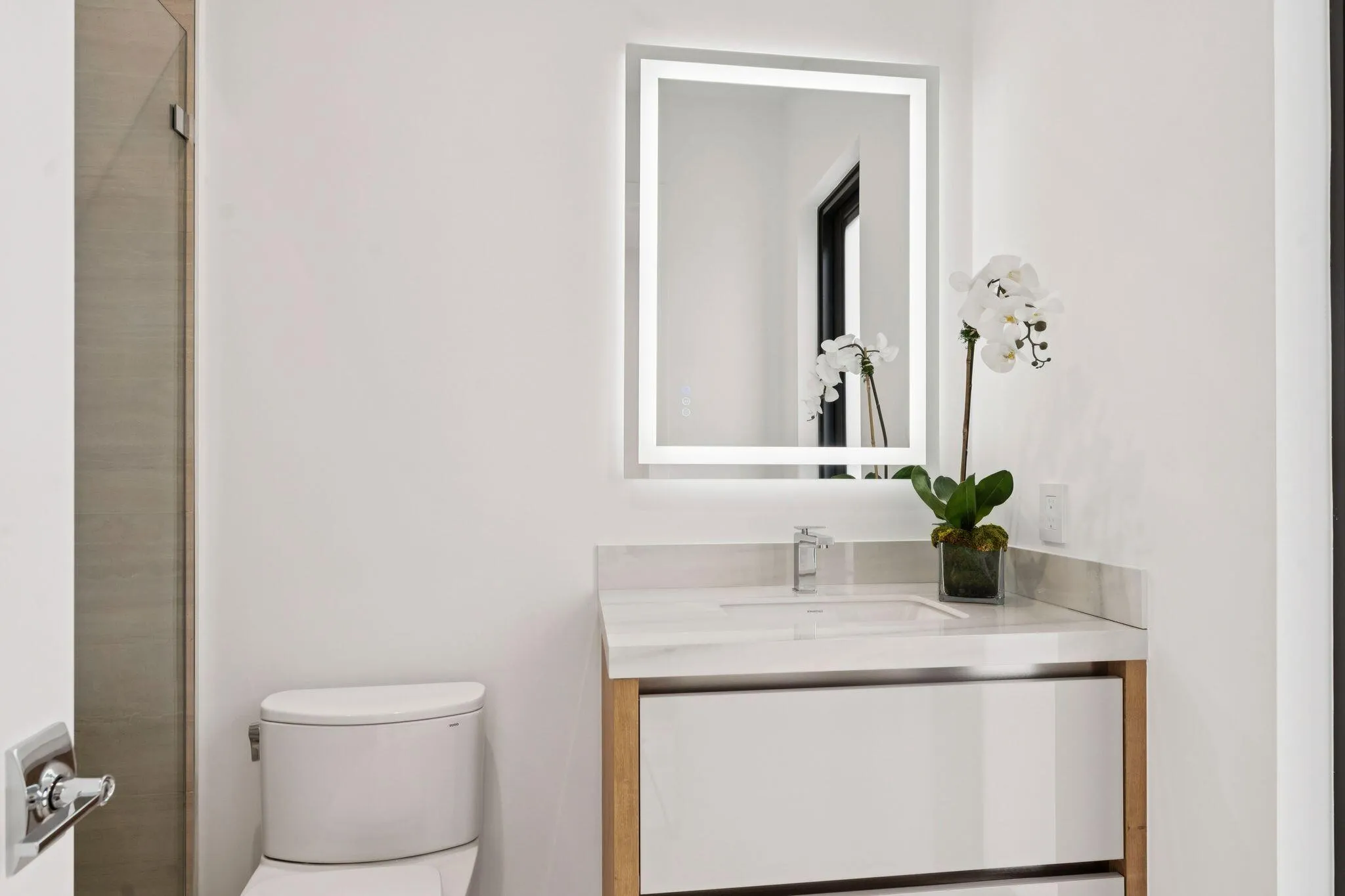 Backlit vanity mirror, single basin sink, white stone countertop, wood-finish vanity with white drawer fronts, and a modern toilet