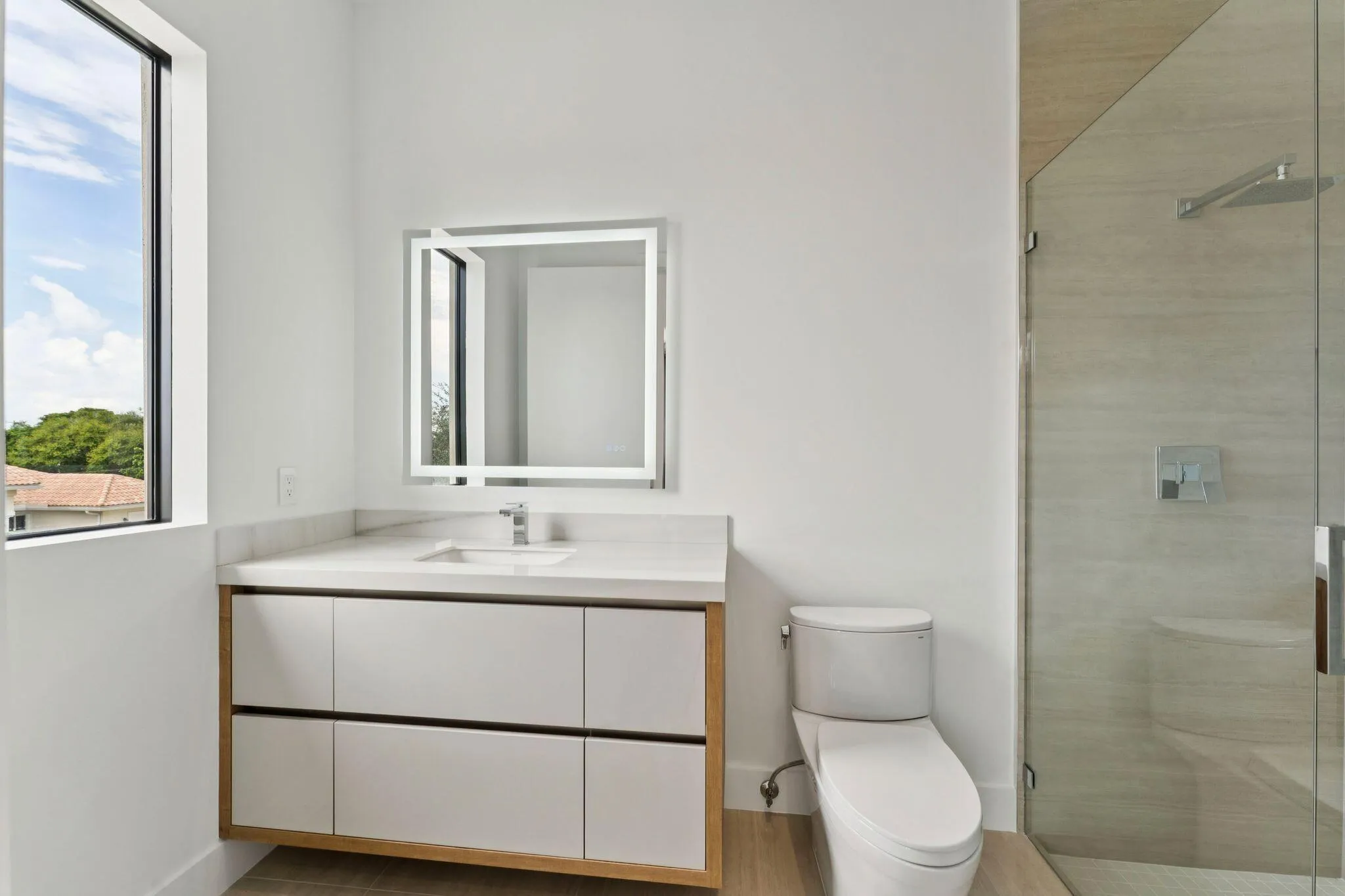 Modern primary bathroom featuring a floating vanity with white cabinetry and wood-finish base, quartz countertop, illuminated mirror with integrated lighting, chrome faucet, porcelain toilet, and a frameless glass-enclosed shower with tile surround and rainfall showerhead