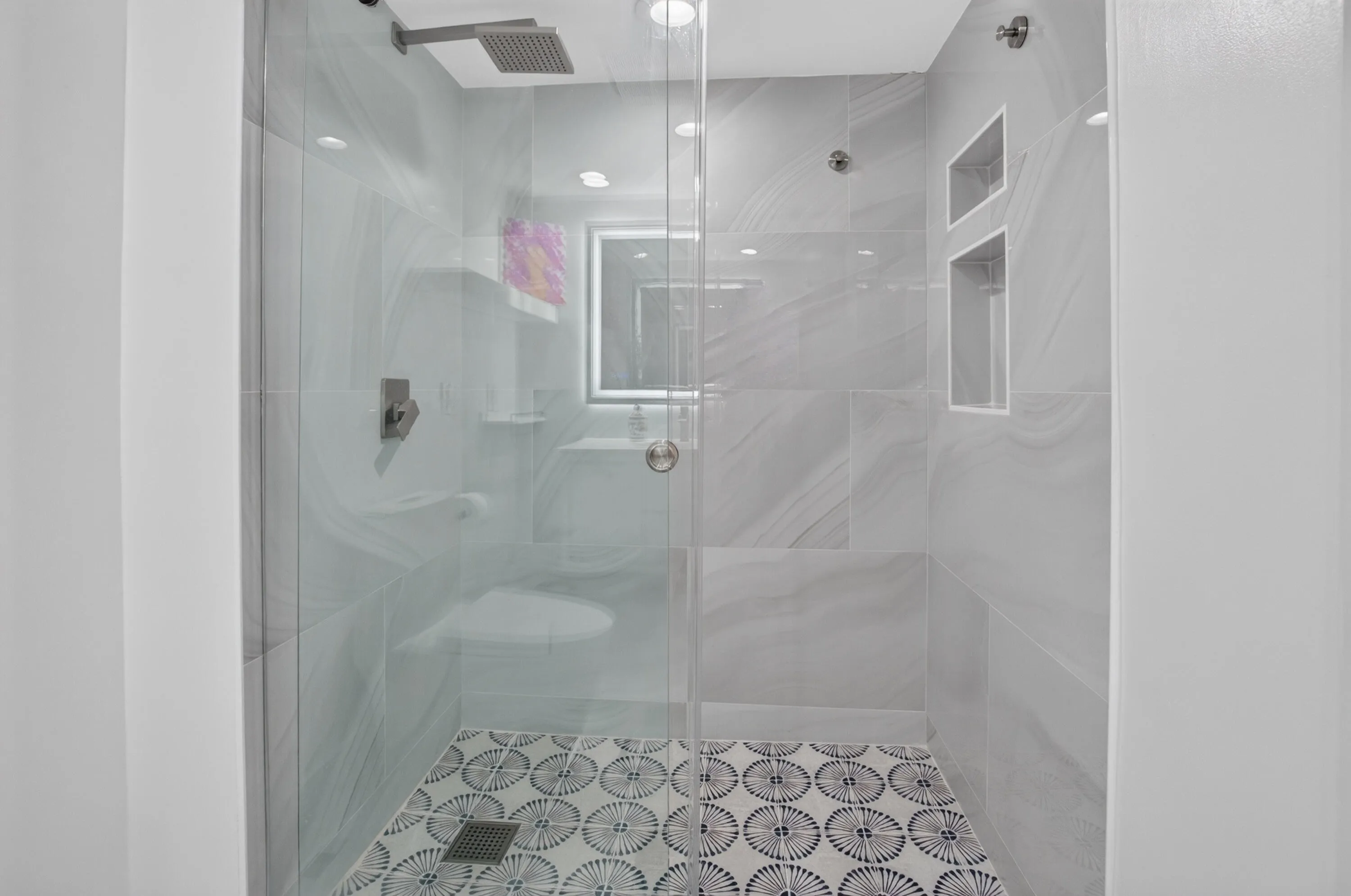 Glass-enclosed shower featuring a rainfall showerhead, large format wall tiles, decorative floor tiles, and recessed shelving