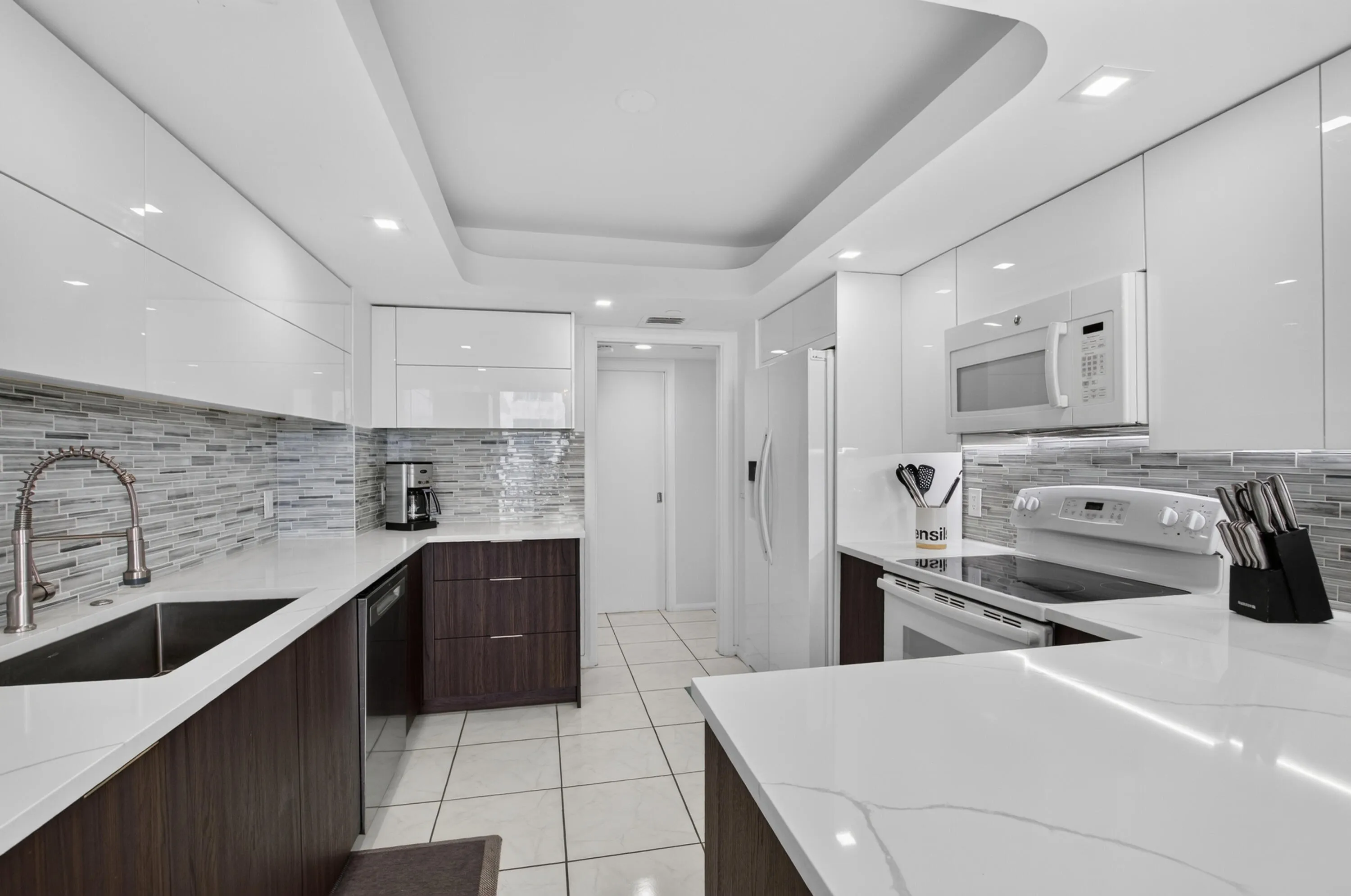 Sleek kitchen featuring white upper cabinetry, dark wood-finish lower cabinetry, white countertops, a mosaic tile backsplash, and recessed lighting