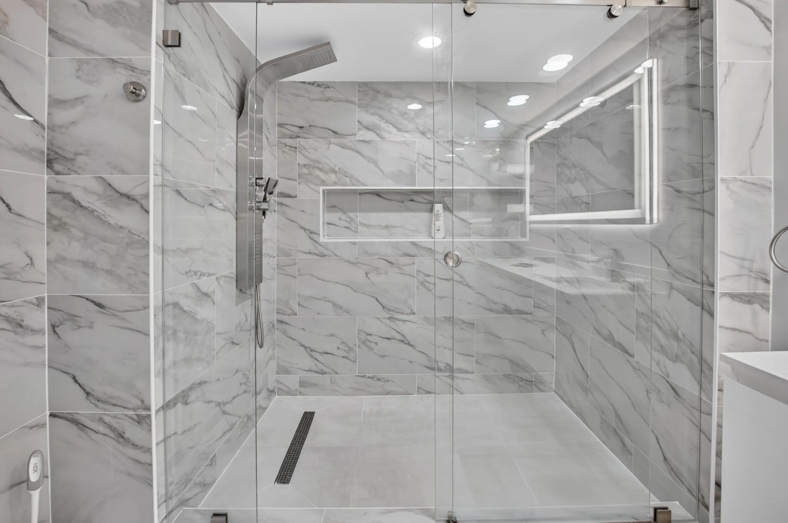Glass-enclosed shower with full-height marble-look tile, linear drain, recessed lighting, and a built-in shower niche