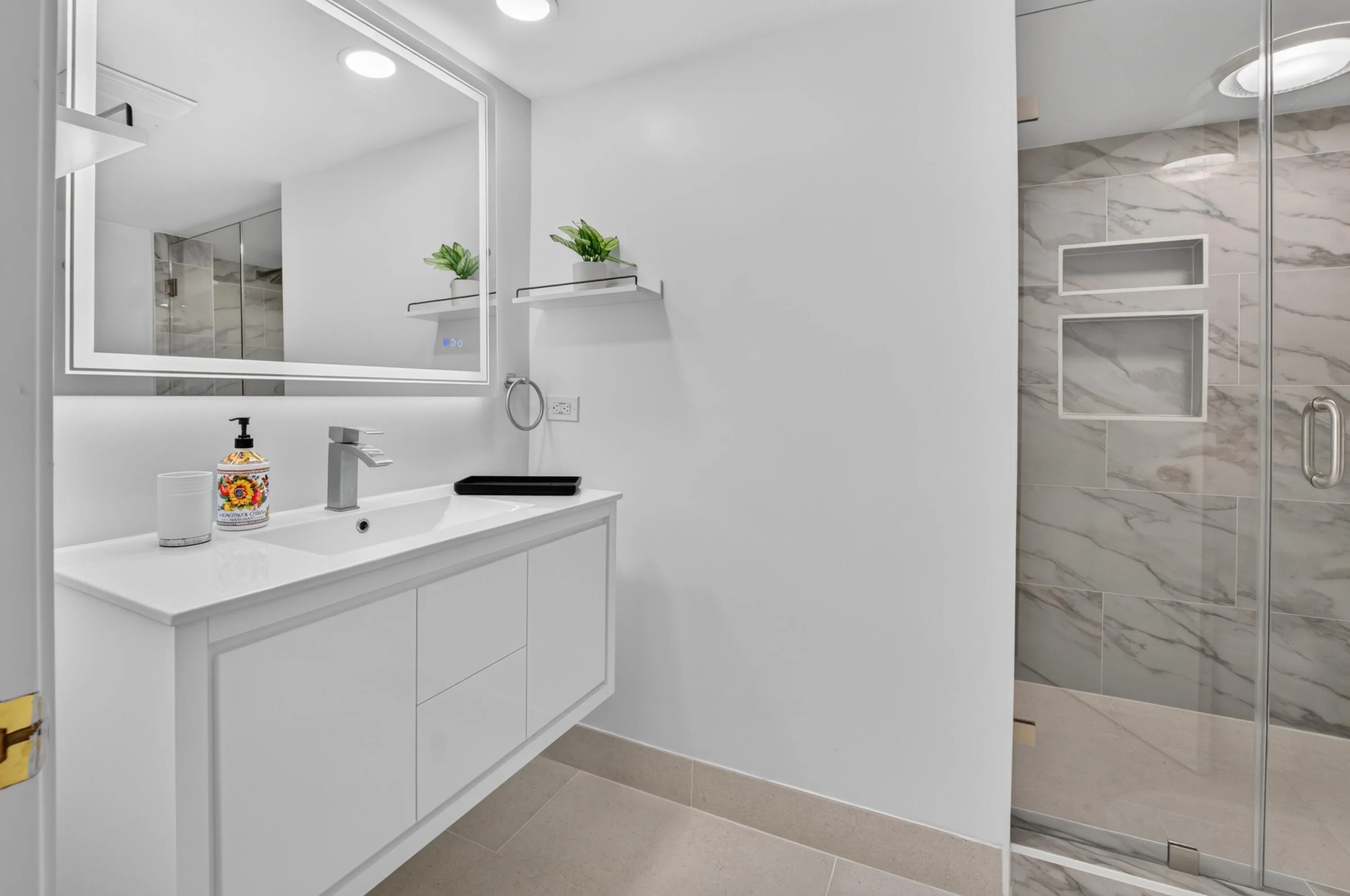 Floating vanity with integrated sink, illuminated mirror, walk-in shower with glass enclosure, marble-patterned tile, and recessed shower niches